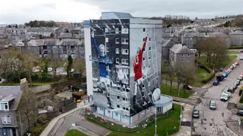 High-rise building with murals of late footballer Denis Law with his arm aloft, one in a red shirt, one in a blue Scotland shirt.