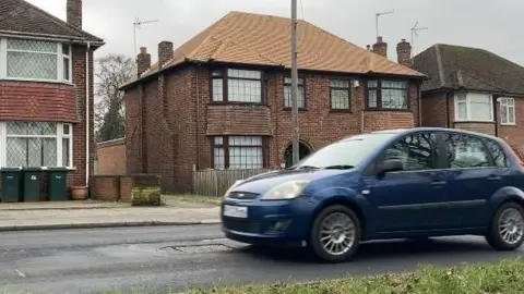 A dark blue car drives over a manhole cover on a residential street - past a number of houses in the background.