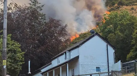 A white two-storey house on a hill is pictured with flames running down the hill towards it. The flames are within metres of the white structure. 