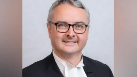 Office of the Clerk of Tynwald Juan Watterson, who has short grey hair and is wearing glasses.