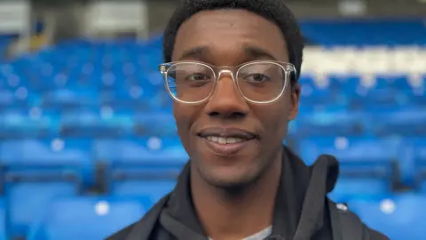 Emma Baugh/BBC Silvio who has neat short black hair and is wearing clear framed glasses and a black hoody. He is starting to smile. Behind him are rows of blue stadium seats. 