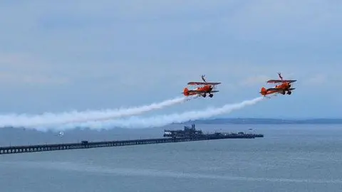 William/Geograph Two red stunt planes flying side by side above the sea in Southend, with a trail of grey smoke behind them. The city's pier is pictured in the background, stretching out to sea.