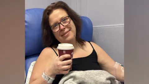 Supplied A woman holds a coffee cup and looks at the camera. She is wearing a black strappy top and has a grey blanket on her lap. She is sitting in a hospital chair.