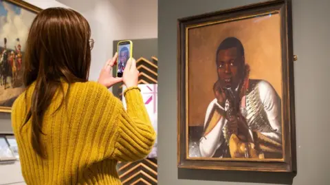 National Army Museum A woman wearing a yellow jumper takes a photo of a painting. The work depicts a black military bandsman holding a cymbal.
