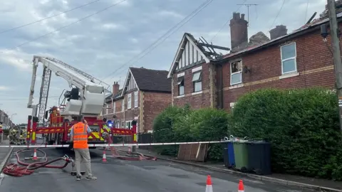 Two fire engines, one with a large crane activated, sit outside a row of terraced houses.  The area on the street has been taped off.  From ground level, you can see the roofs have been burnt away and the upstairs windows burnt and one is blown out.