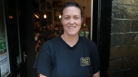 A woman with brown hair slicked back is smiling and wearing a black t shirt which says Roly's Fudge in yellow lettering on it. She is stood in the doorway of a shop.