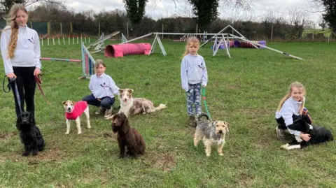 Four children of various ages stand in a dog agility park with a mix of different dogs on leashes. In the background, tunnels, jumps and other obstacles can be seen. 
