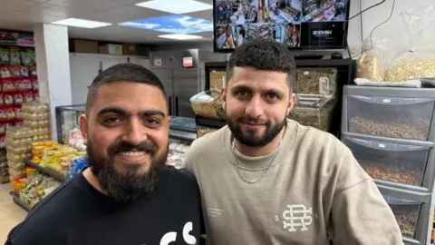 Two men, one in a dark top and one in a lighter top, are standing side by side and smiling. They are in a shop and there are a variety of items in the background.