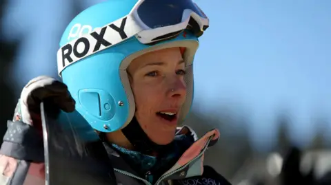 Lesley McKenna is wearing a sky blue helmet with white-rimmed goggles strapped to the top of the helmet. She is holding the top of a snowboard and is looking off to the distance.