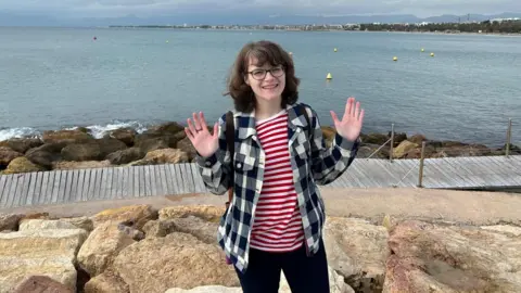 Louise Wallace A woman standing on a rocky outcropping near the sea. She has shoulder length brunette hair, glasses and is wearing a chequed jacket over a red and white striped shirt.