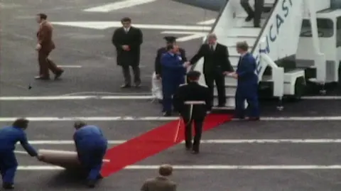 Former president Jimmy Carter coming down the steps from a flight into Newcastle. A red carpet is being quickly rolled out in front of him.