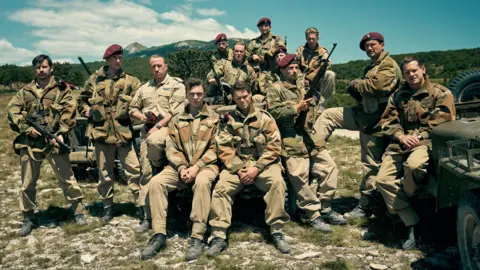 BBC/Baniz Rights/Robert Viglaski a picture of artists of SAS Rogi Heroes. Thirteen men wearing army uniform and purple caps. They are posing with guns on military vehicles and in the hilly surroundings.