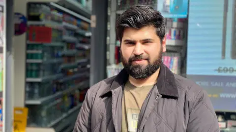 Faisal Umar has dark hair and a beard and is standing outside a convenience store. He is wearing a dark quilted jacket over a brown T-shirt. Shelves stocked with drinks and other products are visible inside the shop behind him.