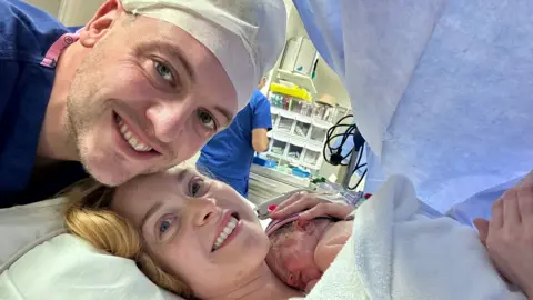 Handout The couple are smiling at the camera in a hospital theatre. Hayley is holding her newborn child.