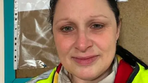 Annabelle Storm has dark hair tied back. She is standing in front of a pinboard and is wearing a high vis jacket over a red top.