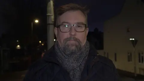 A man with a beard, black glasses, and short dark hair. He is wearing a coat and a grey scarf.