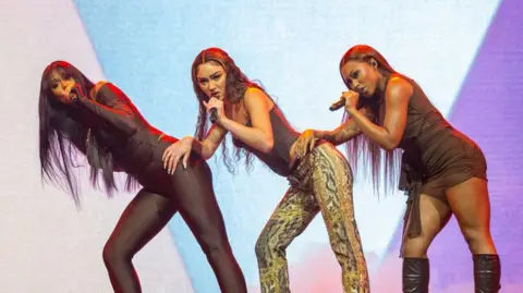 Getty Images  Flo performing on stage.  Jorja Douglas is to the left in a black skin tight catsuit. Stella Quaresma is on the middle in a brown top and animal print trousers. Renee Downer is on the right in a brown dress and black boots.