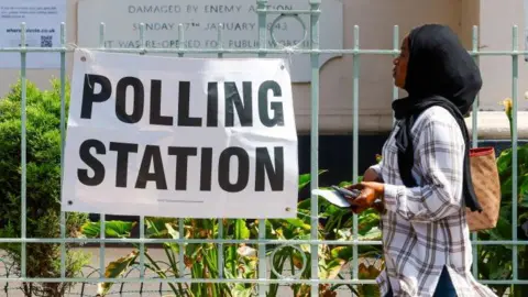 Getty Images A young woman wearing a headscarf walks past a polling station on 4 July 2024. She has a brown shoulder bag and holds a mobile phone in her hand.