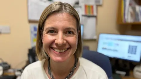 Dr Davies is sat with her desk in the background. She is wearing a white jumper and metallic necklace. She has chin-length fair hair and is smiling at the camera.