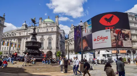 People are seen walking in Piccadilly Circus and sitting on the steps by the fountain. 