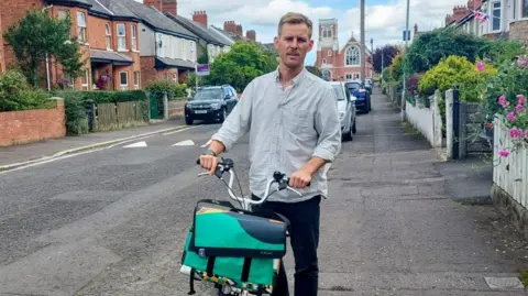 Andrew McClean standing next to his bike on a residential street. He has light brown hair, wearing a light blue shirt and black trousers and watch. 