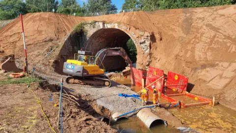 Gary Essex A yellow digger is working next to an arch bridge. There is a brook that is brown and has red construction equipment inside it. The embankment above and around the bridge is brown