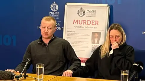 Jordan's stepfather, Michael Groome on the left, sitting behind a table with a glass of water in front of him. He is wearing a dark three-quarter length zip up jumper. His mother, Lynn Gray, is on the left. She has blonde hair and is wearing a dark hoodie. She is holding a tissue to her face with her left arm.