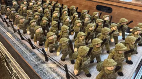An army of woollen soldiers, part of an exhibition of knitted scenes. They are in a glass display case.