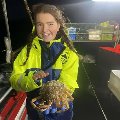 James Robertson Brooke wears a neon yellow and blue waterproof jacket. She stands on board a boat and it is very dark with lights on the boat illuminating the space. She holds a brown crab up for the camera.