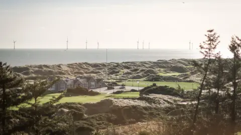 Kris Miller / BBC A golf course with a club house and greens next to the North Sea, which has wind turbines in the distance.