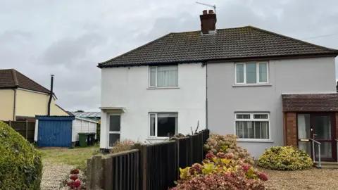 Shaun Whitmore/BBC Semi-detached houses are to the right of the image. The one on the left is painted white and has a garage. On the right is a grey house 