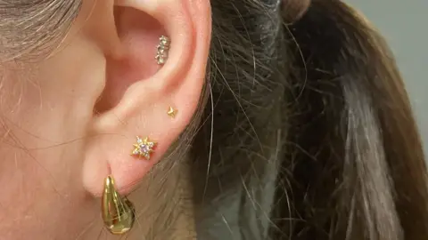 Image shows a close-up of a woman's ear, showing four ear piercings, with gold and diamanté earings, her brown hair tied in a ponytail. 