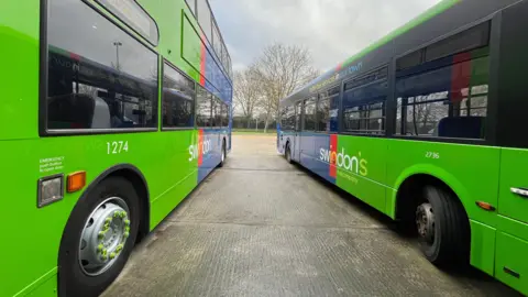 Angle looking down the middle of two buses parked next to each other - green and blue in colour with a Swindon's Bus Company logo.