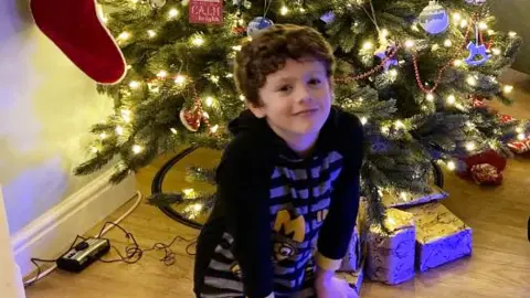 Family photo Dylan Cope who has curly brown hair and is wearing a black hoody with minion characters on it, sitting under a Christmas tree with presents next to him