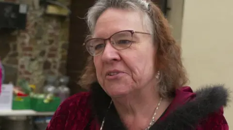 Older woman wearing burgundy framed glasses, with shoulder length wavy hair.  She is wearing ear rings, a neckland and a burgundy, red velvet coat with a a hood edged with black, soft fur.