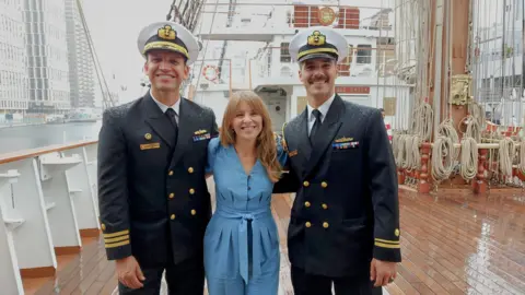 Two naval officers wearing smart uniforms and white military hats stand in the rain either side of a BBC reporter wearing a blue jumpsuit, on the wooden deck of a tallship, with ropes wound neatly behind them 