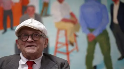Reuters A photo of an elderly gentleman. He wears a white, flat cap, round black glasses, a grey suit jacket and red tie. He sits in front of a large painting or mural which features various men in conversation or standing around in what looks like a bar or other meeting space.