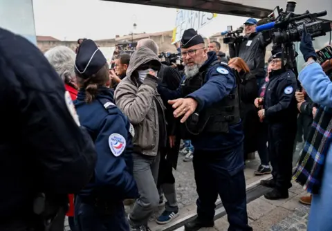 Getty Images Salah satu terdakwa kasus Pelicot yang mengenakan masker, jaket berkerudung, dan kacamata hitam tiba di gedung pengadilan dengan dikelilingi kamera dan polisi.