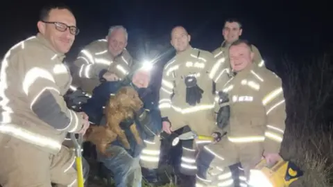 Lincolnshire Fire and Rescue Five firefighters from Lincolnshire Fire and Rescue wearing their uniforms pose for a photo with terrier dog Branston and his owner David Benton. They are smiling at the camera after Branston was rescued.