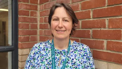 Alison Bell is seen from her shoulders up standing in front of a red brick wall. She has brown hair and is wearing a blue, green and white patterned blouse.
