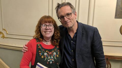 Claire Yates, a woman with red hair and glasses wearing a red top and dungarees, with Dr Michael Mosley