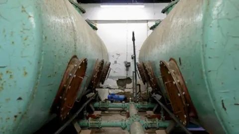 Two large decaying cylinders in a plant room with rusty pipework attached. 
