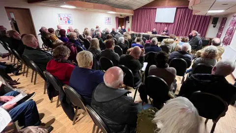 A hall filled with rows of chairs and people sat on them listening to a group of people who are sat at the front of the room. The photo is taken from the back of the room. 