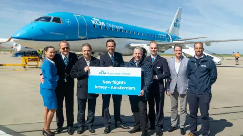 A picture of a number of people stood in a line. A number of people are wearing suits. There is also a sign held up by three people which reads 'New flights Jersey - Amsterdam'. There is a large blue and white KLM plane that they are stood in front of.