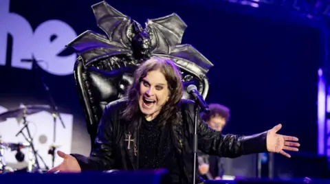 Ozzy Osbourne has long black hair and heavy black eyeliner. He is sitting on a black throne with bat wings on the top, on a stage. He has his arms out and he is singing into a microphone.
