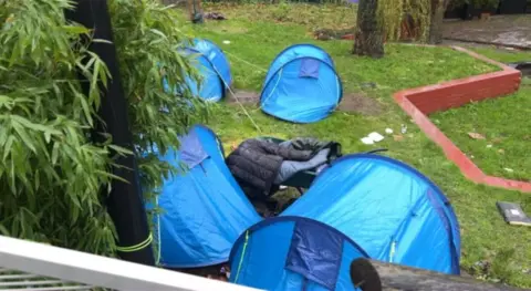 Four blue tents used by rough sleepers in the rain on a patch of grass. 