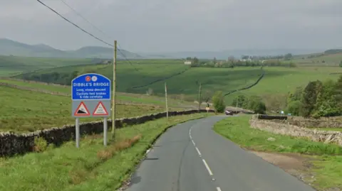 A dales view with a large blue sign that reads 'Dibble's Bridge Long, steep descent. Cyclists test brakes & drive carefully'. 