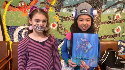 Millie and Joey. Millie has long dark hair, wearing a pink and purple striped top, pink and purple cat ears and face paint giving her a Cheshire cat smile. Joy has long dark hair, wearing a grey beanie hat with felt eyeballs on it. She is holding a Rainbow Fish book.