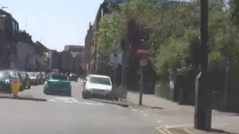 A green car drives down a street lined on either side with parked cars.