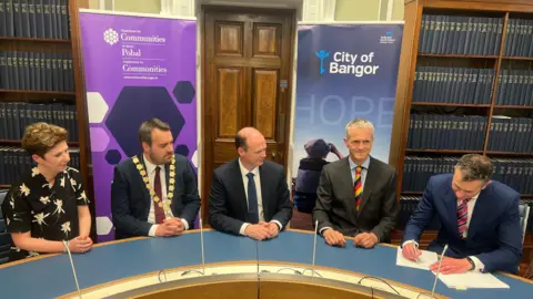 Five people sit along a curved table. There are microphones in front of each one. and two banners behind. A man on the far right is signing a piece of paper.
